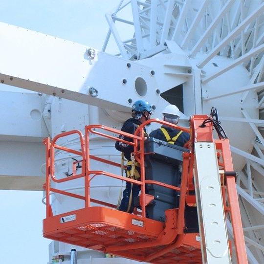 Two men in hardhats standing in an aerial work platform doing antenna maintenance on the back of a ground station